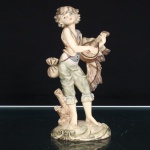 Escultura figurativa em resina policromada, representando jovem músico descalço, em posição ereta, trajando calças arregaçadas, camisa e colete, com pequena bolsa presa à cintura. Sustenta instrumento de cordas dedilhadas de caixa piriforme, alusivo a alaúde ou bandolim, acompanhado por panejamento disposto lateralmente. Composição assentada sobre base oval moldada em relevo, com tronco de árvore integrado à cena. Policromia em tonalidades marfim, verde, cinza e castanho, com pormenores fisionômicos pintados. Iconografia inspirada nas representações europeias de músicos ambulantes e figuras pastoris. Sem marca de manufatura aparente. Medidas: Alt.: 15,5 x Comp.: 9,5 x Larg.: 5,5cm.