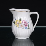 Porcelana Pozzani, Jundiaí - SP (Fundada em 1934 por Francisco Pozzani). Leiteira em porcelana nacional, corpo bojudo moldado em gomos, com colo suavemente estreitado, bico vertedor pronunciado e alça lateral estilizada. Esmaltação branca, decoração floral em estampagem policromada, composta por flores em tonalidades de amarelo, rosa, branco e azul, entre folhagens verdes, concentrada na face principal e repetida em menor escala no reverso. Filete dourado acompanha a borda superior e a alça. Marca de fábrica em verde sob a base: Pozzani -  Jundiaí. Medidas: Alt.: 10 x Comp.: 10 x Larg.: 7,5cm.