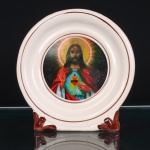 Placa devocional do Sagrado Coração de Jesus executada em faiança, esmaltagem de tom marfim, com suporte integrado à própria estrutura. Ao centro, representação em estampagem policromada do Sagrado Coração de Jesus, com Cristo nimbado, mão direita em gesto de bênção e esquerda indicando o coração flamejante, encimado por cruz e circundado pela coroa de espinhos. Composição delimitada por filetes concêntricos em tonalidade castanho-avermelhada. Base guarnecida por elementos vegetalistas moldados em relevo e esmaltados no mesmo tom. Reverso liso, sem marca de manufatura aparente. Peça de caráter religioso e devocional. Medidas: Alt.: 21 x Comp.: 21 x Larg.: 9cm