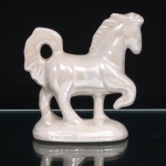 Escultura decorativa em faiança moldada e esmaltada, representando cavalo em atitude de marcha, com uma das patas dianteiras elevada e flexionada. Modelagem estilizada, com crina em relevo, cauda arqueada e vazada, musculatura indicada por sulcos e volumes moldados. Revestimento em esmalte branco de acabamento perolizado e iridescente, produzindo reflexos nacarados conforme a incidência da luz. Assente sobre base oval integral, de contorno irregular. Base inferior oca, com aberturas decorrentes do processo de fabricação. Sem marca de manufatura aparente. Medidas: Alt.: 14,5 x Comp.: 14 x Larg.: 6cm