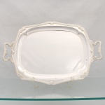 FRACALANZA - MODELO REGENCE. Grande bandeja confeccionada em metal espessurado à prata, c.1930-50, centro ligeiramente rebaixado, borda ondulada contornada por frisos e laços estilizados em relevo, obtidos através da técnica do cinzelado, padrão decorativo replicado nas alças laterais. Marca da manufatura puncionada ao fundo. Medidas: Comp.: x Larg.: cm.