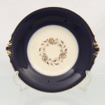 Prato de servir em porcelana alemã KPM, manufatura Krister Porzellan-Manufaktur em Waldenburg, Silésia, período do entre-guerras, circa década de 1930. Centro de fundo marfim ostenta guirlanda circular com três rosas e ramificações fitomórficas transferidas em decalque dourado, aba côncava e profunda apresenta cobertura em tom azul-cobalto esmaltado sob o vidrado, ladeada por duas alças curtas integradas de perfil recortado e relevos em gomos folheados a ouro. Verso apresenta marca carimbada em cinza com coroa imperial, sigla do fabricante, a inscrição "Royal Ivory / A.D. 1831 / Germany" e anotação manuscrita "Echt Kobalt". Possui um fio de cabelo, vide foto. Medida: Diam.: