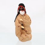 Boneca japonesa (ningyo), representação feminina, confeccionada em estrutura revestida por indumentária têxtil. Figura ajoelhada, trajando quimono em tecido brocado de tonalidade dourada, tecido com motivos florais e elementos geométricos, sobre obi em seda de fundo preto, ornamentado por padrões policromos. Cabeça modelada e policromada, com face em tonalidade branca, olhos, sobrancelhas e boca delineados, olhos semicerrados e boca marcada; cabelo de fibras pretas disposta em penteado tradicional, presa por cordões trançados vermelhos e pequeno adorno em forma de haste. Mãos modeladas, parcialmente inseridas nas mangas, dispostas em gesto frontal. O emprego de tecidos de seda, brocados e fios metálicos integra a tradição da indumentária japonesa documentada em exemplares dos séculos XIX e XX. Medidas: Alt.: x Comp.: x Larg.: cm.