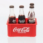 Miniatura promocional Coca-Cola, composto por seis garrafas em vidro, com tampas metálicas e conteúdo líquido, acondicionado em engradado plástico vermelho compartimentado. Garrafas e caixa apresentam inscrições e logotipos "Coca-Cola / Coke  Trade Mark Regd." em branco. Peça de caráter publicitário e colecionável, reproduzindo em escala reduzida as garrafas e o engradado de transporte da marca. Faltando duas garrafas. Estado do conteúdo desconhecido.Medidas caixote: Alt.: 3,5 x Comp.: 8 x Larg.: 5cm.Medidas garrafa: Comp.: 7,8cm.