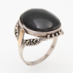 JOIA. Anel confeccionado em prata, com aro sustentando mesa oval elevada, centrada por cabochão de pedra negra, possivelmente ônix. Galeria vazada com elementos lineares e granulação, guarnecida lateralmente por marcassitas. Aplicações retangulares em metal de tonalidade dourada dispostas nos ombros. Superfície com oxidação nos recessos. Trabalho do século XX. Medida: Diam.: 2 cm; Peso bruto: 6.1 gramas.