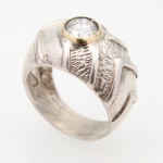 JOIA. Anel masculino confeccionado em prata, de aro largo, com mesa centrada por pedra incolor de lapidação brilhante, engastada em caixa circular de metal dourado com quatro garras. Ombros estruturados por faixas geométricas em relevo, alternando superfícies polidas e campos de textura cinzelada. Medida: Diam.: 2,3 cm. ; Peso: 8 gramas.