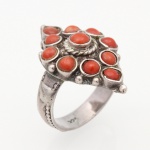 Anel confeccionado em prata, com mesa losangular estruturada em composição simétrica de onze cabochões de coral, dispostos em engastes individuais de aro liso; elemento central elevado, circundado por cercadura de fio torcido. Campo ornamentado por granulação e pequenos motivos aplicados. Ombros e aro cinzelados com elementos fitomórficos e sucessão de discos em relevo. Apresenta punção de garantia da prata do toque 0,925.  Medida: Diam.: 2 cm. ; Peso bruto: 6,3 gramas.