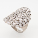 Anel confeccionado em metal prateado, de aro largo, com mesa oval de perfil abaulado e estrutura vazada, inteiramente guarnecida por numerosas pedras incolores de lapidação baguete, cravadas individualmente em disposição irregular e multidirecional, formando composição de caráter geométrico. Galeria inferior vazada, acompanhando a distribuição das cravações. Medida: Diam.: 2 cm; Peso bruto: 9.1 gramas.