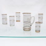 Conjunto para refresco em vidro incolor, circa 19501960. Composto por uma(01) jarra e cinco(05) copos, com decoração de temática náutica. Jarra de corpo cilíndrico, ligeiramente alargado junto à base, bico vertedor e asa aplicada em vidro maciço; copos de paredes retas. Ornamentação constituída por navio à vela, executado em traço dourado, articulado a filetes horizontais concêntricos, com frisos da mesma tonalidade junto às bordas e bases. Medidas: jarra: Alt.: 23,5 x Comp.: 17 x Larg.: 11 cm; Copo: Alt.: 12,5 x Diam.: 7 cm.