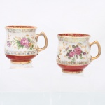 Duas(02) canecas comemorativas em porcelana. Corpos de perfil abaulado sobre base circular, com asas arqueadas e superfície revestida por esmalte nacarado de efeito lustre. Decoração policroma por decalcomania com ramagens de rosas, complementada por arabescos e frisos florais em ouro. Faixa inferior e interior da boca em esmalte vermelho, esta última ornamentada por motivos fitomórficos dourados. Reservas posteriores com a inscrição "Felicidades" em caligrafia dourada. Bordas, asas e pés realçados a ouro. Medidas: Alt.: 9 x Comp.: 10,5 x Larg.: 8,5 cm.