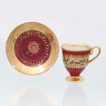 Polovi Comércio de Porcelanas Ltda, Campo Largo - PR. Chávena para café em porcelana nacional. Revestimento em esmalte de tonalidade vermelho e faixas em douração, apresentando friso central com cena equestre em silhueta, composta por cavaleiros, vegetação e construções. Barrados ornamentais dourados delimitam a composição, com friso geométrico no interior da borda. Pires circular com campo central vermelho, medalhão em forma de estrela e sucessão de motivos vegetalistas, circundado por aba integralmente dourada com ornamentação floral. Marca de manufatura "Polovi - Paraná - Made in Brazil" no reverso. Medidas: Xícara: Alt.: 7 x Comp.: 8 x Larg.: 6cm; Pires: Diam.: 10,5 cm.