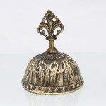 Sineta de mesa em bronze, corpo campaniforme fundido, com ornamentação em relevo composta por aves afrontadas entre elementos vegetalistas e friso perimetral. Empunhadura elevada, vazada, em forma de palmeta estilizada, assentada sobre colar moldurado. Interior com badalo metálico suspenso. Medidas: Alt.: 11,5 x Diam.: 9,5 cm.