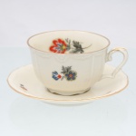 Xícara para chá com pires em porcelana, manufatura KAHLA, Alemanha, circa 1930-1940. Corpo moldado em porcelana de tonalidade marfim, com discretos relevos florais junto à borda e asa em voluta. Decoração policroma por decalcomania, composta por motivos florais estilizados em vermelho, azul, amarelo e grisaille, distribuídos no bojo, interior e pires, realçado por filetes dourados nos contornos. Marca de fábrica no reverso do pires: escudo coroado com inscrição "KAHLA" e Padrão "Forma Livia". Medidas: Xícara: Alt.: 5 x Comp.: 10,5 x Larg.: 8,5 cm; Pires: Diam.: 13 cm.