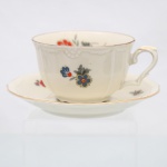 Xícara para chá com pires em porcelana, manufatura KAHLA, Alemanha, circa 1930-1940. Corpo moldado em porcelana de tonalidade marfim, com discretos relevos florais junto à borda e asa em voluta. Decoração policroma por decalcomania, composta por motivos florais estilizados em vermelho, azul, amarelo e grisaille, distribuídos no bojo, interior e pires, realçado por filetes dourados nos contornos. Marca de fábrica no reverso do pires: escudo coroado com inscrição "KAHLA" e Padrão "Forma Livia". Medidas: Xícara: Alt.: 5 x Comp.: 10,5 x Larg.: 8,5 cm; Pires: Diam.: 13 cm.