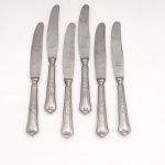 HERCULES INOX. Modelo Classique. Conjunto composto por seis(06) facas médias confeccionadas em aço inoxidável, datável do período circa 1960-1970. Executados ao estilo francês do período Luís XV, apresentam cabos de perfil sinuoso e terminação espatulada, enriquecidos por ornamentação seriada em relevo que conjuga motivos de volutas, rocalhas e filetes perimetrais. Lâminas exibem gume com acabamento serrilhado de fábrica e faces frontais com gravação em matriz ácida "Hercules AÇO INOX", integrada à efígie do fabricante sob o selo de têmpera especial "GELODUR". O reverso exibe a marca em baixo-relevo "HERCULES INOX". Medidas: Comp.: 21 cm.