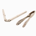 Dois(02) quebra-nozes em metal prateado, modelos de acionamento manual: um em forma de tenaz, com braços alongados e mordentes internos canelados para apreensão e ruptura da casca; outro articulado em alavanca, com cabos cilíndricos texturizados e mecanismo serrilhado junto à articulação. Apresentam sinais de uso, oxidação e desgaste do prateado.Medidas: Comp.: 15,5 cm; Comp.: 12,5 cm.