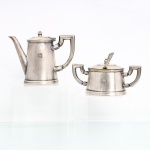 Serviço para café em metal prateado, manufatura Fracalanza. Composto por cafeteira e açucareiro, ambos de corpo cônico, base oval moldurada e tampas articuladas. Cafeteira provida de bico alongado e alça angular; açucareiro com duas alças laterais simétricas e colher própria. Corpos ornamentados por monograma geométrico "H" gravado. No reverso, punções da manufatura Fracalanza, acompanhadas de numeração de fabricação. Apresentam sinais de uso e desgaste do prateamento. Medidas bule: Alt.: 12,5 x Comp.: 16,5 x Larg.: 7cm; Medidas açucareiro: Alt.: 8,5 x Comp.: 17 x Larg.: 8cm; Medida colher: Comp.: 13,5 x Larg.: 3cm.