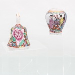 Dois(02) itens em porcelana: um sino(falta badalo) de mesa e um minivaso, decoradosm ao estilo Família Rosa (Famille Rose), ornamentação contínua do tipo Rose Canton pintada em esmalte sobre o vidrado, caracterizada por grandes corolas de peônias centrais em matizes rosa-choque, circundadas por densa ramagem de folhagens verdes, flores secundárias azuladas e borboletas dispersas sobre fundo esmaltado ocre, o vaso com acabamento em filete dourado na orla, bojo com duas cartelas opostas delimitadas por molduras retangulares pretas em padrão Rose Medallion, sendo que a reserva principal abriga cena palaciana narrativa com três figuras tradicionais em trajes civis de época ladeadas por mobiliário e janelas abertas, enquanto a reserva oposta ostenta motivos fitomórficos com flores e frutos. Medidas sino: Alt.: 7,5 x Diam.: 5 cm; Medidas vaso: Alt.: 6 x Diam.: 5 cm.