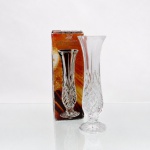 Vaso solitário(solifleur / bud vase) em cristal de chumbo a 24% (24% Lead Crystal),produzido pela marca francesa Cristal d'Arques (J.G. Durand) na França, pertencente à linha decorativa Masquerade. Apresenta formato de trompete com bocal expandido e corpo alongado que afunila em direção à base, assentando-se sobre pé circular plano decorado com sulcos radiais na parte inferior. O terço inferior do corpo recebe ornamentação moldada com padrão facetado em cortes cruzados e pontas de diamante geométricos, encimados por orlas em formato de leque e gomos lisos verticais que acompanham a curvatura superior do gargalo. Acompanha caixa cartonada original. Medidas: Alt.: 18 x Diam.: 5,5 cm.