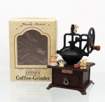 Caixa de música mecânica executado em forma de moedor de café, comercializado sob a denominação "Antique Coffee-Grinder", de fabricação japonesa. Apresenta estrutura confeccionada em material plástico imitando madeira e metal, composta por base quadrangular com gaveta frontal amovível dotada de puxador metálico em relevo. A seção superior ostenta engrenagens simuladas e uma grande roda lateral de manobra mecânica com cabo de madeira torneada. A composição cênica é integrada por três figuras tridimensionais em resina ou plástico moldado representando camundongos. Possui mecanismo de corda inferior, o sistema reproduz uma melodia enquanto executa movimentos simulados sincronizados na roda e nas personagens (simulated action). Acompanha caixa cartonada original de fábrica com ilustrações de cariz nostálgico e inscrições comerciais "Playing Musical / Antique Coffee-Grinder". Medidas: Alt.: 18 x Comp.: 11,5 x Larg.: 10,5 cm.