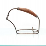 Suporte para garrafa de vinho (cesta ou berço de serviço) confeccionado em metal e madeira. A peça apresenta estrutura aramada simplificada com base elíptica de perfil baixo projetada para a acomodação e inclinação da garrafa. A seção traseira dispõe de gancho vertical em formato de "L" para travamento do fundo do vasilhame, enquanto a seção frontal eleva-se em curva sinuosa terminando em um anel circular fechado destinado à contenção e passagem do gargalo; alça superior arqueada recebe pega anatômica central em madeira torneada, fixada à armação de metal por duas ponteiras cônicas. Medidas: Alt.: 20,5 x Comp.: 25 x Larg.: 8,5cm.