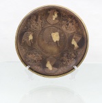 Prato decorativo de parede em latão, decorado com temática egípcia, centro brunido em tonalidade castanha e decoração incisa com detalhes em metal aparente. Composição organizada em medalhões, tendo representação de faraó em perfil, circundada por retratos e cenas inspiradas na iconografia do Antigo Egito, incluindo figura identificável como Nefertiti, pirâmides, esfinge, carruagem, personagens e inscrições hieroglíficas de caráter ornamental. Bordo delimitado por faixa dourada. Reverso com argola metálica para suspensão. Apresenta oxidação e marcas de uso. Medidas: Diam.: 19,5 cm.