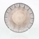 Cesto fruteiro confeccionado em metal prateado, formato circular de perfil baixo, com fundo plano e aba vazada por sequência radial de recortes oblongos, formando padrão regular ao redor do centro. Bordo recortado em arcos sucessivos e apoio sobre pequenos pés esféricos. Superfície com oxidação, escurecimento e sinais de uso. Medidas: Diam.: