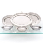 Conjunto de seis(06) peças para servir, confeccionado em metal prateado. Composto por três travessas ovais de diferentes dimensões, uma delas com bordo recortado e moldurado por frisos, e três pequenas tigelas individuais de formato circular, com abas laterais. Superfícies lisas, sem ornamentação, com sinais de uso e oxidação compatíveis com o material. Medidas bandeja maior: Alt.: 2 x Comp.: 35,5 x Larg.: 25,5cm.Medidas bandeja média: Alt.: 2 x Comp.: 30 x Larg.: 21cm.Medidas bandeja menor: Alt.: 2 x Comp.: 25 x Larg.: 18cm.Medidas consomé: Alt.: 3,5 x Comp.: 17 x Larg.: 14,5cm.