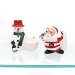 Par de esculturas natalinas em cerâmica esmaltada, representando Papai Noel e boneco de neve, modeladas em cerâmica e policromadas em vermelho, verde, preto e tons rosados sobre fundo branco. Cada figura incorpora recipiente aberto na parte posterior, destinado ao acondicionamento de pequenos objetos, guloseimas ou composição de mesa natalina. Decoração aplicada sob esmalte vítreo, com detalhes do vestuário e atributos característicos das figuras. Medidas maior: Alt.: 14 x Comp.: 14 x Larg.: 8 cm; Medidas menor: Alt.: 11,5 x Comp.: 14 x Larg.: 11 cm.