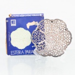 Esteira para mesa em metal prateado, manufatura Eberle, fabricação nacional. Peça de formato circular, constituída por placa vazada de contorno lobulado, com composição simétrica de rosáceas e volutas estilizadas distribuídas radialmente. Estrutura elevada sobre pequenos apoios cilíndricos, destinada à proteção da superfície da mesa contra o contato direto com recipientes de serviço. Acompanha embalagem original da Eberle, com a inscrição "Esteira para Mesa". Apresenta sinais de oxidação e desgaste do revestimento metálico. Medidas: Alt.: 1,5 x Comp.: 14,5 x Larg.: 13,5 cm.
