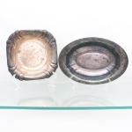 Duas(02) travessas em metal prateado. Composto por exemplar oval e outro quadrangular de cantos arredondados. Uma apresenta fundo oval rebaixado, aba compartimentada por nervuras radiais e faixa perimetral ornamentada em relevo com volutas, rosetas e motivos fitomórficos, a outra possui fundo circular rebaixado e quatro reservas côncavas dispostas junto às bordas, contorno moldurado e perfil recortado. Necessitam polimento e/ou banho de prata. Medidas: Alt.: 4,5 x Comp.: 22,5 x Larg.: 22,5 cm; Medidas: Alt.: 4 x Comp.: 30 x Larg.: 20 cm.