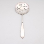 Colher para servir tomates ou frutas confeccionada em metal espessurado a prata, manufatura estadunidense International Silver Company em Meriden, Connecticut, datável da primeira metade do século XX. Apresenta cabo longo de perfil plano com linhas puras e extremidade arredondada. A concha circular e côncava destaca-se pelo trabalho vazado central e simétrico que reproduz ramos de videira compostos por folhas e frutos estilizados (tomates ou bagas) dispostos em torno de um medalhão circular liso, configuração técnica destinada a permitir o escoamento de líquidos durante o serviço de mesa. O reverso do cabo ostenta três punções gravadas em baixo-relevo: a chancela institucional "INTERNATIONAL SILVER CO.", o monograma heráldico de garantia "IS" inserido em molduras quadradas individuais e a marca de origem de linha "ORIGINAL ROGERS". Medidas: Comp.: 19 x Larg.: 7cm.