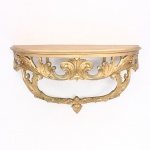 Console de parede em estilo Luís XV, construído em madeira cmo revestimento de pátina dourada., tampo semicircular, circundado por saia perimetral recortada e ricamente ornamentada por volutas entrelaçadas, concheados(rocaille) e folhas de acanto vazadas em alto-relevo. Assente sobre duas pernas de perfil sinuoso em curva e contracurva, convergentes na porção inferior em um vértice decorado por pingente escultórico em formato de pinha ou cacho de frutos estilizados. O reverso revela o chassi de fixação estrutural provido de cantoneiras metálicas. Medidas: Alt.: x Comp.: x Larg.: