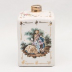 Frasco de toucador em porcelana, executado ao gosto rococó. Corpo paralelepipédico de cantos arredondados, colo cilíndrico e bocal com aplicação dourada. Superfície esmaltada em branco, decorada por decalcomania policroma com cena, representando casal em jardim junto a elemento arquitetônico; faces laterais e ombros com ramos florais. Composição delimitada por filetagem e elementos geométricos a ouro. Fundo com marca pintada, não identificada. Manufatura não determinada, circa 1950-1970. Apresenta desgaste pontual da douração; tampa não acompanha. Medidas: Alt.:13,5 x Comp.: 8,5 x Larg. 8,5cm.