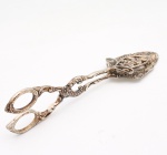 Pegador ou tesoura de servir para confeitaria confeccionado em metal espessurado a prata, de fabricação europeia ou norte-americana, datável de meados do século XX. Apresenta estrutura articulada por rebite central ocultado por florão em relevo de ambos os lados. Os braços de manobra exibem empunhadura em formato de tesoura com argolas ovais ornamentadas por friso perolado e terminação em ponta folheada. As hastes centrais são trabalhadas com motivos vazados e volutas acantiformes em relevo pronunciado. A extremidade funcional dispõe de duas garras assimétricas destinadas ao manuseio de doces: a seção inferior configura uma espátula de contorno recortado e perfil plano com recortes vazados lineares, enquanto a seção superior adota formato lanceolado em estilo neo-renascentista, inteiramente decorada por arabescos, folhagens e flores integradas à estrutura calada. Apresenta oxidação e marcas de uso. Medida: Comp.: 22cm.