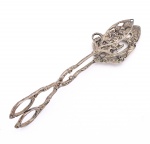 Pinça para servir doces confeccionada em metal prateado. Corpo articulado em forma de tesoura, com decoração vazada e relevada de motivos florais e folhagens. Pegadores ovais, hastes ornamentadas e articulação central em roseta. Extremidades de preensão largas, destinadas ao serviço de bombons, petits fours, biscoitos e outras peças de confeitaria. Apresenta oxidação e sinais de uso. Medida: Comp.: