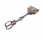 Pinça para servir bolo confeccionada em metal prateado. Estrutura articulada em forma de tesoura, com empunhaduras anelares e hastes ornamentadas por motivos florais em relevo. Extremidades providas de duas pás retangulares vazadas, com contorno recortado e composição de rosas, flores e folhagens em relevo, destinadas à apreensão e ao serviço de doces, petits fours e peças de confeitaria. Superfície com oxidação e desgaste do prateamento. Medidas: Comp.: 18,5cm.