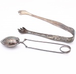 Dois(02) itens em metal prateado: uma pinça para açúcar e um infusor de chá. Pinça de braços arqueados, hastes alargadas e extremidades recortadas em forma de garras, destinada à apreensão de cubos de açúcar. Infusor constituído por cápsula ovoide perfurada, formada por duas calotas articuladas, acoplada a mecanismo de abertura por hastes e empunhadura em anel. Superfícies com sinais de oxidação e desgaste do revestimento metálico. Medidas pinça: Comp.: 15cm.Medidas infusor: Comp.: 17cm.