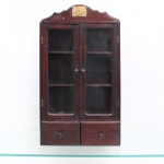 Armarinho vitrine de parede(cristaleira suspensa de pequenas dimensões), executado pela Southwood Factory, circa 1950. Estrutura de formato retangular confeccionada em madeira de tonalidade tabaco com veios aparentes, coroada por frontão superior de perfil recortado em ondulações simétricas; porção frontal dispõe de duas portas envidraçadas com puxadores esféricos torneados em madeira, que dão acesso ao espaço interno compartimentado por duas prateleiras, gerando três nichos de exposição; base do móvel integra dois gaveteiros paralelos providos de puxadores idênticos aos das portas. Topo do frontão ostenta placa retangular em latão fixada por parafusos fenda, trazendo a inscrição da marca gravada em baixo-relevo: "SOUTHWOOD / FACTORY". O reverso plano em madeira preserva duas alças metálicas circulares superiores para suspensão. Estyado conforme fotografias. Medidas: Alt.: 68,5 x Comp.: 38,5 x Larg.: 12 cm.