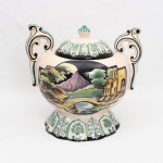 Grande vaso decorativo com tampa(potiche) executado em faiança, de procedência portuguesa, produzido nas oficinas cerâmicas de Alcobaça, circa 1950. Apresenta corpo globular bojudo flanqueado por duas alças laterais elevadas com terminação em volutas recurvadas de caráter rococó, assentando sobre base circular alteada em pedestal de contorno lobado. Ornamentado por duas cartelas pictóricas principais em reservas quadrangulares sobre fundo preto: a primeira exibe uma cena de gênero bucólica ou romântica com duas figuras femininas em trajes tradicionais inseridas em paisagem arborizada com edificação ao fundo; a segunda ilustra um cenário arquitetônico com ponte arqueada sobre curso d'água e torre fortificada de caráter historicista. Tampa removível de perfil hemisférico coroada por pomo em formato de botão estriado, exibindo motivos fitomórficos radiados em reserva. Decoração policromada sob o vidrado em tons de verde-cobre, ocre, azul e vináceo. Verso da base ostenta inscrição caligráfica manuscrita pelo artesão: "Alcobaça / Pintado à Mão / 459", acompanhada pelo monograma ou assinatura de oficina. Medidas: Alt.: x Comp.: x Larg.: