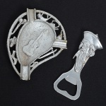 Dois(02) itens: a) Cinzeiro em metal prateado, estrutura vazada com elementos decorativos diversos e reserva central em forma de violão em alto-relevo. Formato irregular, com três tubos laterais funcionais como descanso de cigarro. Medidas: Alt.: 11 x Comp.: 8 cm. b) Abridor de garrafas em metal, com cabo moldado em formato de figura masculina com barba longa e gorro estilizado, em relevo. Boca do abridor integrada ao corpo da peça. Medida: Comp. 10,5 cm.