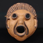 Máscara cerâmica em terracota modelada, possivelmente de teatro grego, de formato antropomórfico, apresentando olhos circulares vazados em relevo, nariz saliente, boca circular aberta e decoração linear pintada em tonalidade escura sobre fundo terroso. Provida de cordão para suspensão.  Medidas: Alt.: 30 x Larg.: 26,5