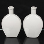Duas(02) garrafas executadas em porcelana, esmaltagem vítrea na cor branca, bojo achatado de seção oval, gargalo cilíndrico e curto, borda ligeiramente ressaltada, apresenta perda da tampa. Med.: Alt. 20 x Comp. 12,5 x Larg. 6 cm.