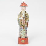 Escultura executada em porcelana chinesa moldada e pintada, representando Shou Xing (também denominado Shou Lao), divindade taoísta associada à longevidade. Apresenta-se pé sobre base retangular, portando bastão cerimonial e colar de contas (chaozhu). Veste túnica longa esmaltada em policromia no estilo Famille Rose, com decoração de faixas verticais, nuvens e motivos simbólicos, usa chapéu cônico vermelho com ponta central e possui barba aplicada em relevo, base com reserva floral pintada nas faces. Período Republicano (19121949). Fundo contendo marca não esmaltada, típica de oficinas da região de Jingdezhen. Med.: Alt. 25 x Comp. 7 x Larg. 6 cm.