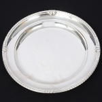 WOLFF. Grande bandeja circular confeccionada em metal espessurado à prata, centro ligeiramente rebaixado, borda contornada por barrado relevado com padrão geométrico em repetição, intercalado por folhagens e volutas estilizadas. Marca da manufatura puncionada no verso da borda. Apresenta marcas de uso. Med.: Alt. 3 x Diam. 33