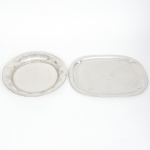 Duas(02) bandejas confeccionadas em aço inoxidável, uma de formato retangular e bordas arredondas, outra circular relevada com folhagens estilizadas. Medidas: Alt.: 3,5 x Diam. 36 cm; Comp.: 43,5 x Larg.: 33,5 cm.