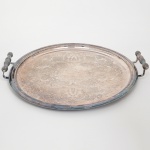 Bandeja circular confeccionada em metal espessurado à prata, centro ligeiramente rebaixado, lavrado com composição de motivo vegetalista, borda de contorno relevado, alças laterais fusiformes, cobertas por pétalas e friso perlado, apoiadas sobre haste curvada. Necessita polimento. Medidas: Alt.: 6,5 x Comp.: 48 x Larg.: 44,5 cm.