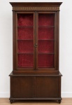 Palaciana vitrine em madeira com duas portas envidraçadas, quatro prateleiras de vidro, interior revestido em veludo vermelho com marcas do tempo, friso superior em bronze dourado e armário inferior removível com duas portas e prateleira interna. Medidas: 196 x 104 x 42 cm (alt. x larg. x prof.).