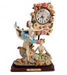 Relógio de mesa De Elina, em resina italiana policromada, representando duas bailarinas entre flores, com base em madeira e mecanismo de quartzo a pilha. Apresenta leves sinais do tempo e pequeno desgaste no vestido de uma das figuras, conforme fotos. Sem garantia funcionamento. Medidas: 29 x 18 cm.
