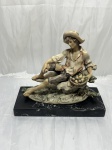 Escultura em resina policromada representando jovem camponês com chapéu assentado sobre rocha ao lado de cesta de frutas, fixada sobre base retangular de mármore preto. Medindo base 27cm x 11,5cm x 15cm de altura total.